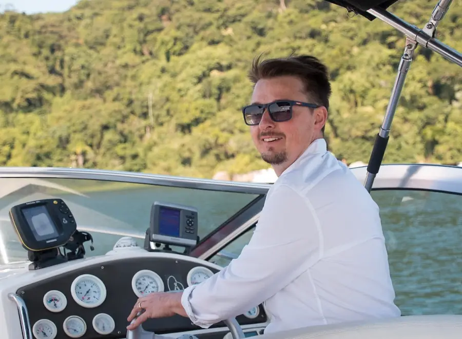 Tiago Mance à la barre d'un bateau à moteur, en chemise blanche et lunettes de soleil, la main sur le volant, devant deux écrans de navigation et les cadrans du tableau de bord.