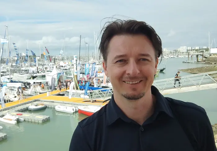 Tiago Mance, souriant, en chemise sombre, devant un port de plaisance rempli de bateaux, des tentes blanches au loin.
