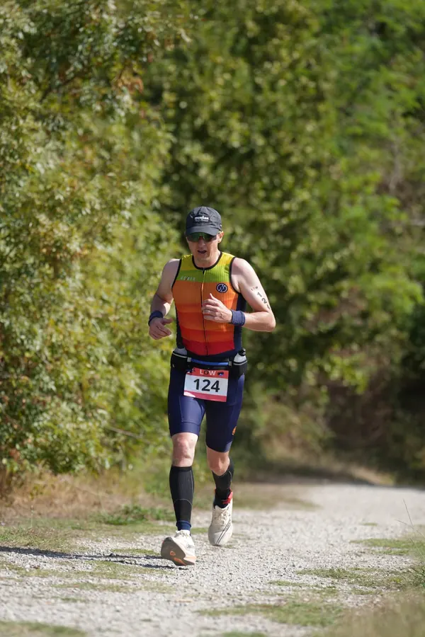 Course à pied, de loin : Tiago Mance, en tenue orange et jaune, casquette et dossard, court sur un chemin de gravier bordé d'arbres.